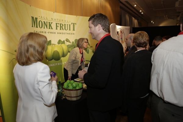 Several attendees gather at the Monk Fruit booth. A man in a dark jacket and a woman in a white jacket face the table, while a woman in a tan jacket stands behind it near a basket of green fruit.
