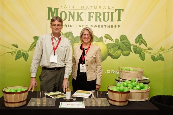A man and a woman, both wearing red lanyards and badges, stand behind a table at the Monk Fruit booth. Baskets of green fruit, glasses, a bottle and brochures sit in front of a green Monk Fruit banner.