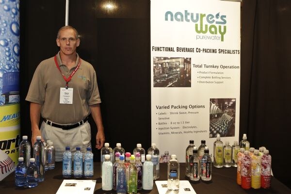 A man in a tan polo with a red lanyard stands behind a black-draped table of bottled waters, including Essentia, next to a Nature's Way Purewater co-packing banner.