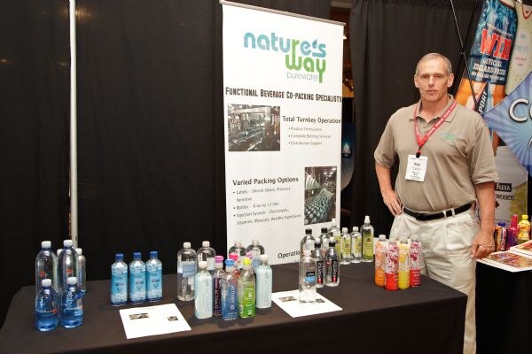 A man in a tan polo with a red lanyard stands beside a table of bottled waters, including Essentia, and a Nature's Way Purewater co-packing banner.