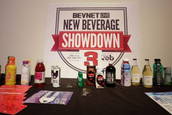 A table in front of a BevNET Live New Beverage Showdown 3 sign holds bottles, cans and cartons, including Cold Brew, Sipp, Power Coco and omega water, with flyers.