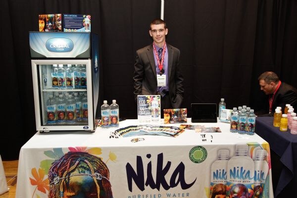 A young man in a dark suit with a lanyard stands behind a Nika Purified Water table. A Crystal Cooler holds Nika bottles beside him, and a seated man is at right.