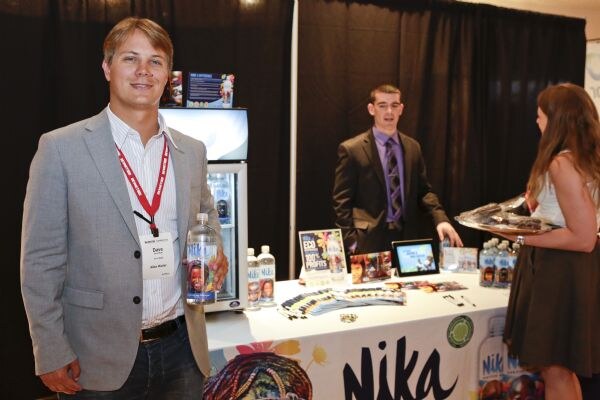 A man in a gray blazer with a red lanyard holds a Nika water bottle beside a booth. Behind the Nika table a man in a suit stands near a cooler, and a woman holds a tray.