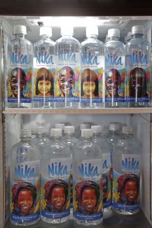 Two shelves of Nika Purified Water bottles in a lit cooler, each label showing a smiling child's portrait against a colorful floral background.