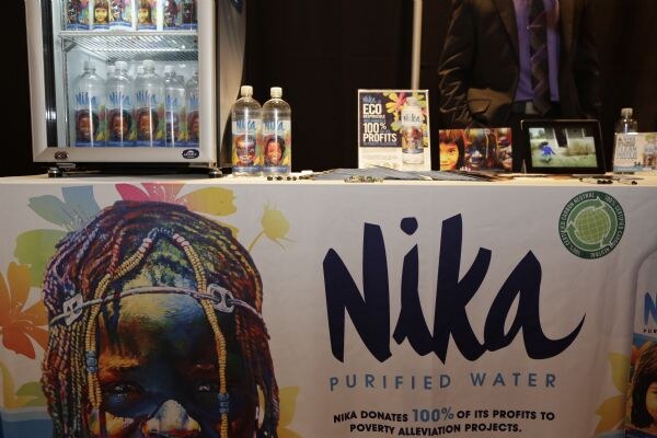 A table cover reading Nika Purified Water with a painted portrait. Nika bottles, flyers and a tablet sit on top, with a small cooler behind and a person in a suit partly visible.