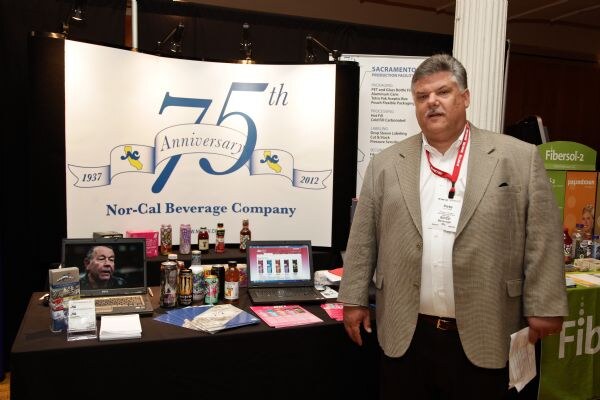 A man in a gray blazer with a red lanyard stands beside a Nor-Cal Beverage Company booth with a 75th Anniversary banner, laptops and bottles on the table. A Fibersol-2 display is at right.