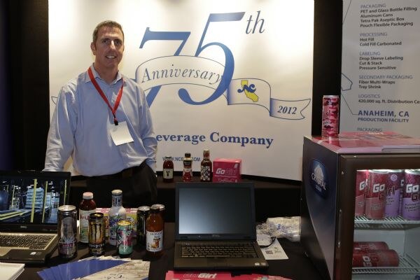 A smiling man in a light blue shirt and red lanyard stands at a table with laptops and canned drinks in front of a 75th Anniversary banner. A cooler of Go Girl cans is at right.