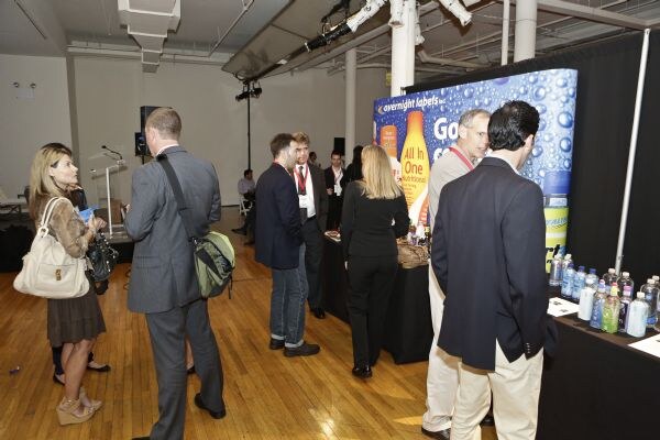 Attendees stand and talk in a loft-style room near an Overnight Labels booth with a bubble-patterned backdrop and bottles on a table. A podium is in the background.