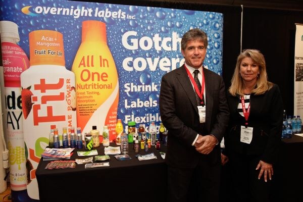 A man in a dark suit and a blonde woman, both wearing red lanyards, stand at the Overnight Labels Inc. booth. A bottle-label backdrop and a table of beverages are behind them.