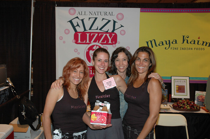 Four smiling women with arms around each other stand in front of an All Natural Fizzy Lizzy banner; one holds a four-pack of grapefruit soda. A Maya Kaimal sign is beside it.