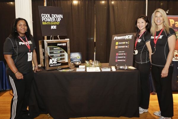 Three women in black PowerIce shirts stand around a draped black table holding a PowerIce display case, signs and sample items.