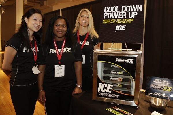 Three smiling women in black PowerIce shirts and red lanyards stand beside a PowerIce display case and a sign reading Cool Down, Power Up.