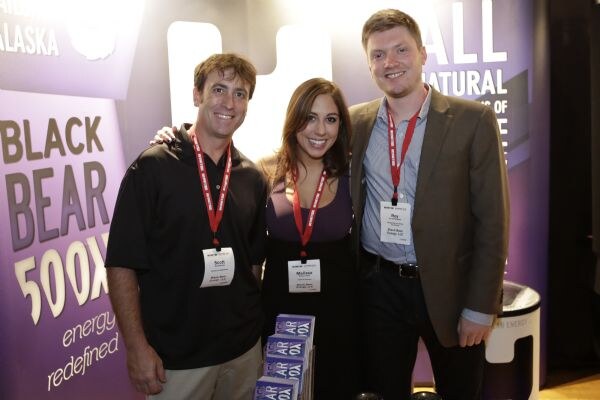 Scott, Melissa and Roy, wearing red lanyards, smile together in front of purple Black Bear 500X signage, with boxes of product on the table in front of them.