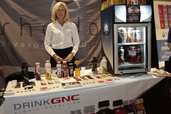 A blonde woman in a white shirt stands behind a table of GNC drinks and printed cards, with a Crystal Cooler refrigerator and a Shadow Beverages banner behind her.