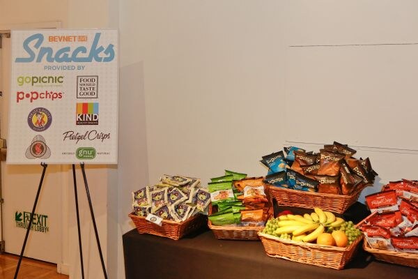 A BevNET Live Snacks sign lists sponsors GoPicnic, Popchips, Food Should Taste Good, KIND, Pretzel Crisps and GNU, next to baskets of snacks and fruit.