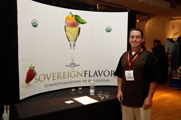 A smiling man in a dark shirt and red lanyard stands beside the Sovereign Flavors booth, which has a curved backdrop with a champagne glass, strawberry and USDA Organic seals, and a table with small cups.