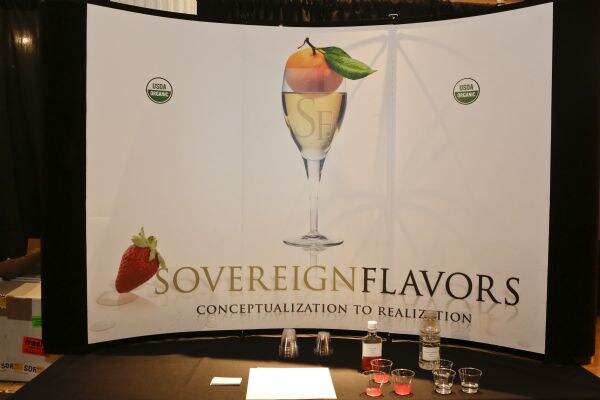 Sovereign Flavors backdrop showing a champagne flute with a peach and a strawberry, above a table with small cups of pink liquid and two bottles.