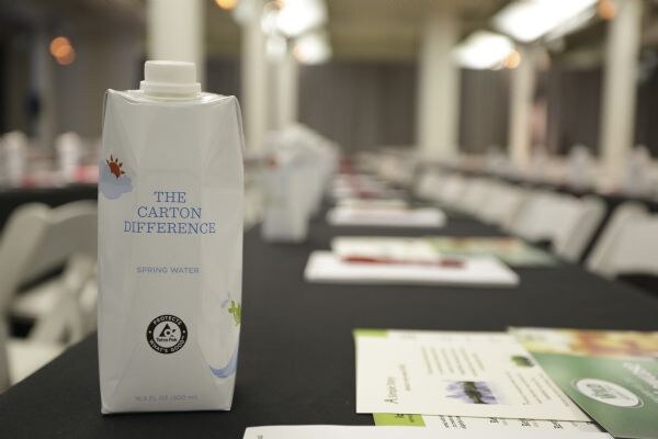 A white Tetra Pak carton labeled The Carton Difference, Spring Water stands on a black-draped table. Brochures lie in front, and rows of tables and chairs blur in the background.