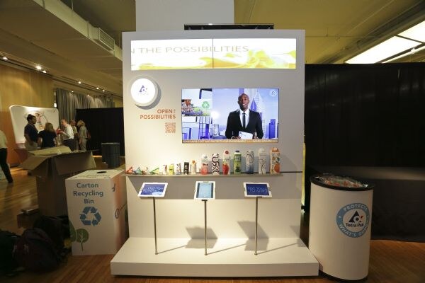 A Tetra Pak exhibit with a video screen of a man in a suit, a shelf of beverage cartons and bottles, three tablets on stands, a carton recycling bin and a round Tetra Pak podium.