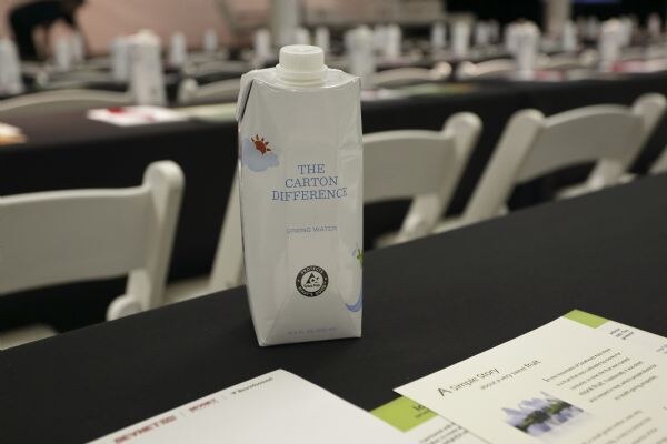 A The Carton Difference spring water carton sits on a black table in front of white folding chairs and printed handouts in a hall.