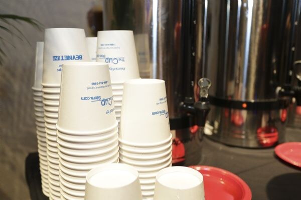 Stacks of white paper cups printed with TheCupStore.com and BevNET Live sit on a table beside two metal beverage urns and red plates.