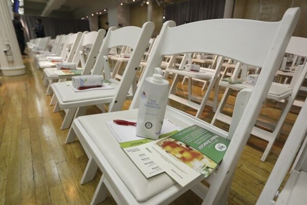White folding chairs in rows hold sponsor materials. In front, a bottle labeled The Gaston Pepperwoods sits on brochures and a red pen, and the chairs behind have wrapped items on their seats.