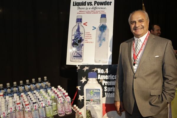 A smiling man in a gray suit and red lanyard stands beside a VBlast Liquid vs. Powder poster and a table of bottled water bottles.