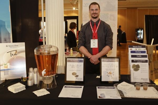 A bearded man in a dark shirt and red tie stands behind a table with a Virginia Dare tea dispenser, sample cups, signs and brochures.
