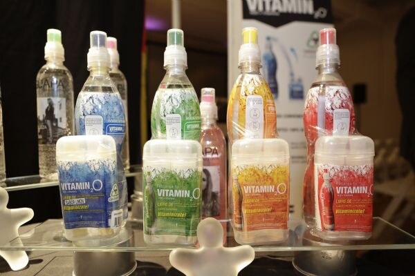 Vitamin O2 packages in blue, green, yellow and red stand on a glass shelf in front of sport-cap bottles with matching colors. A Vitamin O2 banner is in the background.