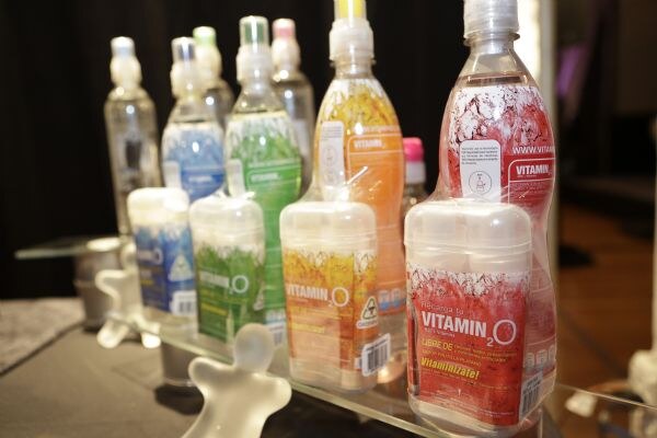 Side view of Vitamin O2 packages in blue, green, orange and red lined up on a glass shelf in front of colored bottles.
