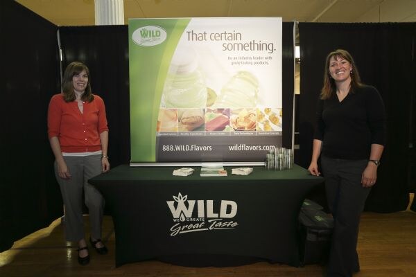 Two smiling women stand on either side of a Wild Flavors table with a banner reading That certain something against black curtains.