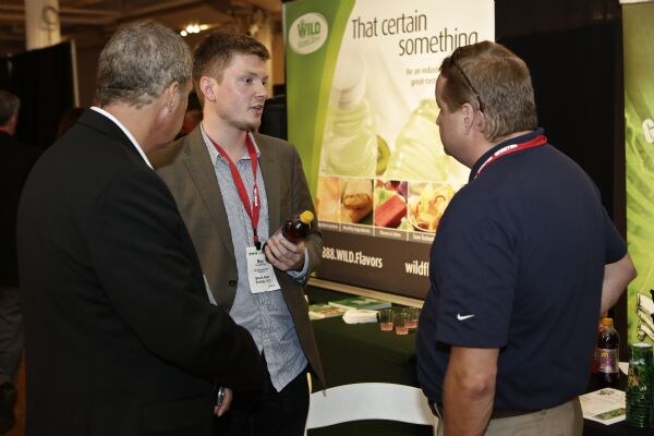 Three men talk at a Wild Flavors booth with a "That certain something" banner. One holds a bottle, and another wears a Nike polo.