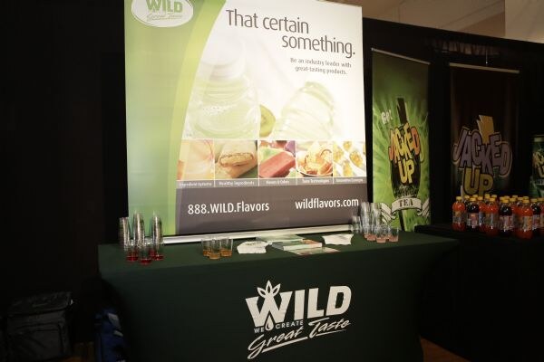 A table with a green Wild Flavors cover and a banner reading That certain something, with sample cups. Jacked Up banners and bottles stand to the right.
