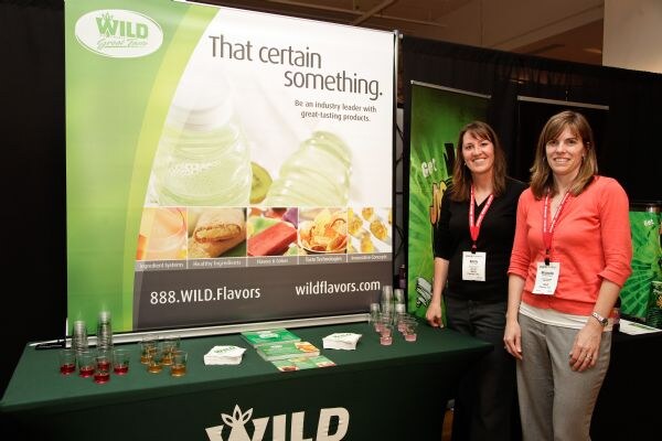 Two women with red lanyards stand beside a Wild Flavors table with a That certain something banner and sample cups.