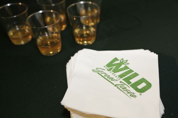Four small clear cups of amber liquid sit on a dark tablecloth beside a stack of white napkins printed with the green Wild logo.