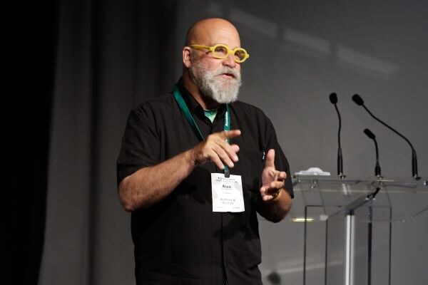 Alan of Alchemy & Science, a bald man with a gray beard and yellow glasses, wears a black shirt and lanyard and gestures with both hands while speaking near a podium with microphones.