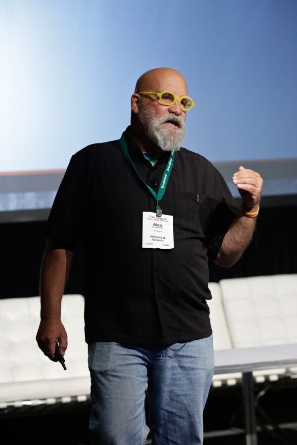 Alan of Alchemy & Science, a bald man with a gray beard and yellow round glasses, wears a black shirt and green lanyard badge and gestures while speaking on stage in front of white leather couches.