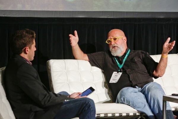 Alan of Alchemy & Science, with yellow glasses and a gray beard, sits on a white leather couch with both hands raised, facing a man in a dark jacket on the left who holds a tablet.