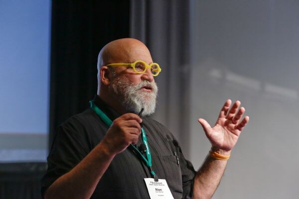 Alan, a bald man with a gray beard and yellow round glasses, wears a dark shirt and green badge lanyard and raises one open hand while speaking, holding a small clicker in the other hand.