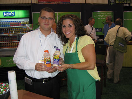 A man in a white shirt and glasses and a woman in a green apron hold Naked juice bottles together at a booth with Naked signs and a refrigerated display behind them.