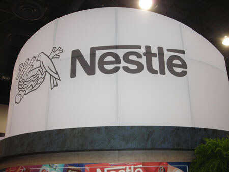 Large white curved sign reading Nestlé in gray letters beside the Nestlé bird-and-nest logo, above a booth counter with partial Nestlé product graphics at the bottom edge.
