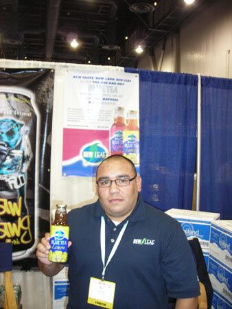 A man with glasses and a dark New Leaf polo holds a bottle of New Leaf Blue Tea Lemon in front of a New Leaf poster, with a Raw Dawg banner at the left and boxes behind him.