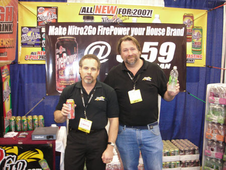 Two men in black polos with lanyards stand at a booth, one holding a FirePower can and the other a bottle, under a banner reading Make Nitro2Go FirePower your House Brand.