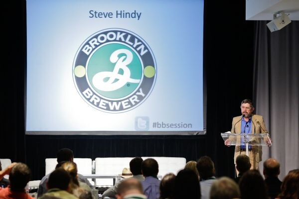 A man in a tan jacket speaks at a clear podium beside a large screen showing the Brooklyn Brewery logo, the name Steve Hindy and the hashtag #bbsession, with audience members seen from behind.