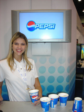 A smiling woman with long blonde hair and a lanyard stands at a counter with Pepsi cups, in front of a wall-mounted screen showing the Pepsi logo.