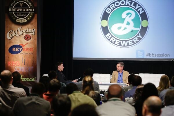 Two men sit on white couches on stage, one in a tan jacket and one in a dark jacket, beneath a screen with the Brooklyn Brewery logo. A banner with sponsor logos stands left, and the audience is in front.