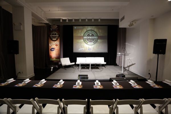 A conference room with a stage, white seating, a podium and a projected Brewbound logo. A sponsor banner including Key Keg stands at left. Rows of tables with brochures face the stage.