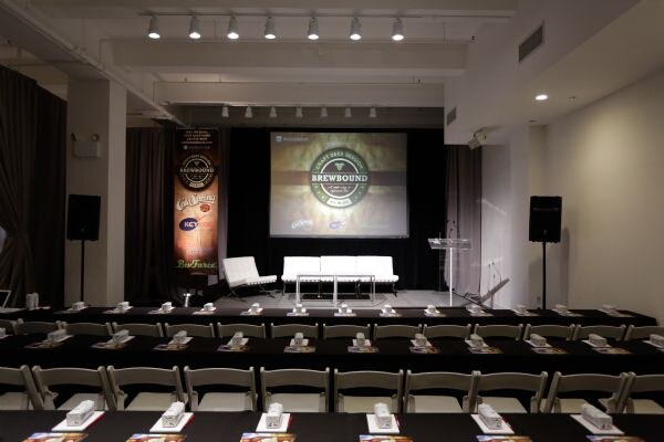 View from the back of a conference room toward a stage with a projected Brewbound logo, white chairs and a podium. A banner with the Key Keg logo stands at left, and tables and folding chairs fill the foreground.