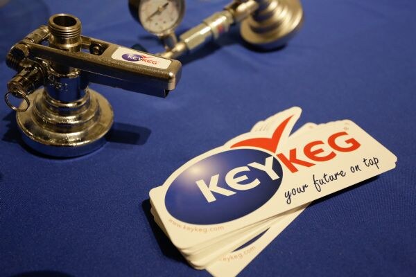 KeyKeg stickers reading "your future on tap" lie on a blue tablecloth beside a metal keg coupler with a KeyKeg handle label and a pressure gauge.