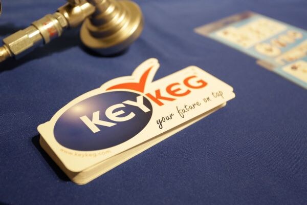 A stack of KeyKeg stickers reading "your future on tap" sits on a blue tablecloth next to a metal keg coupler and a printed card.